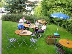 Gäste sitzen an runden Tischen im sonnigen Hotelgarten unter einem Sonnenschirm